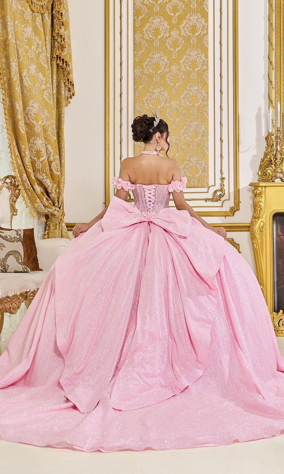 A woman wearing the Quinceanera Dress 5115 By Dancing Queen, a light pink ballgown with a large bow, sits facing away in an ornate room adorned with gold accents and patterned drapes.